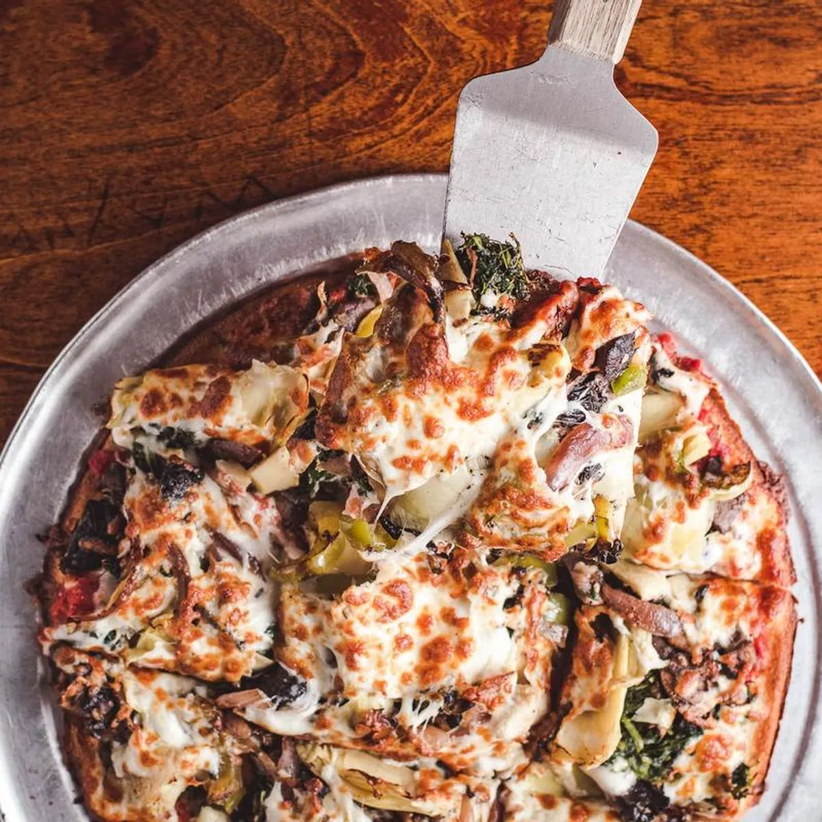 Overhead of a loaded pizza on a steel pan, artichoke, spinach and mushroom under browned cheese