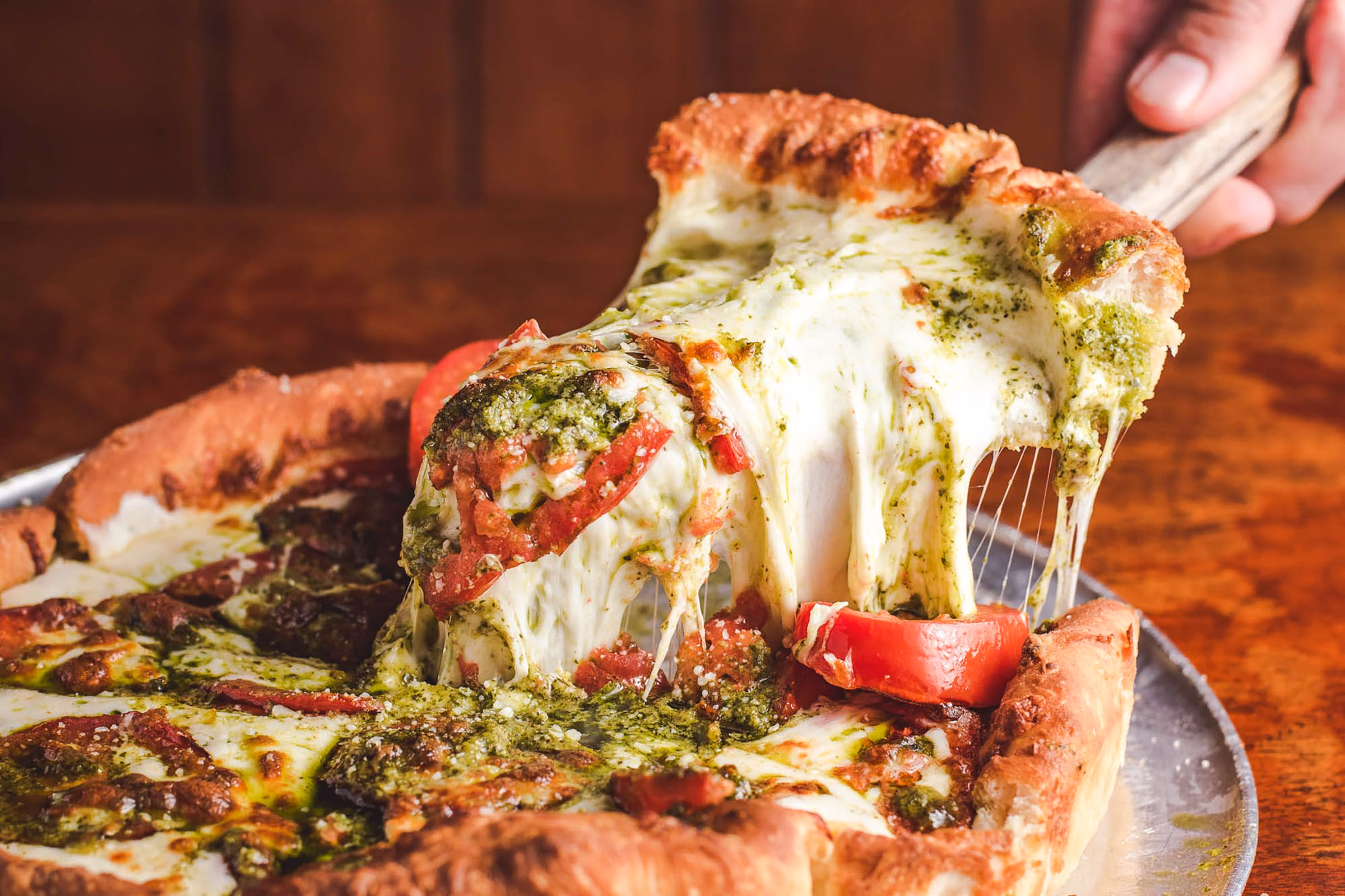 A slice of stuffed deep dish lifted from the pan on a spatula, cheese stretching, pesto and tomato on top