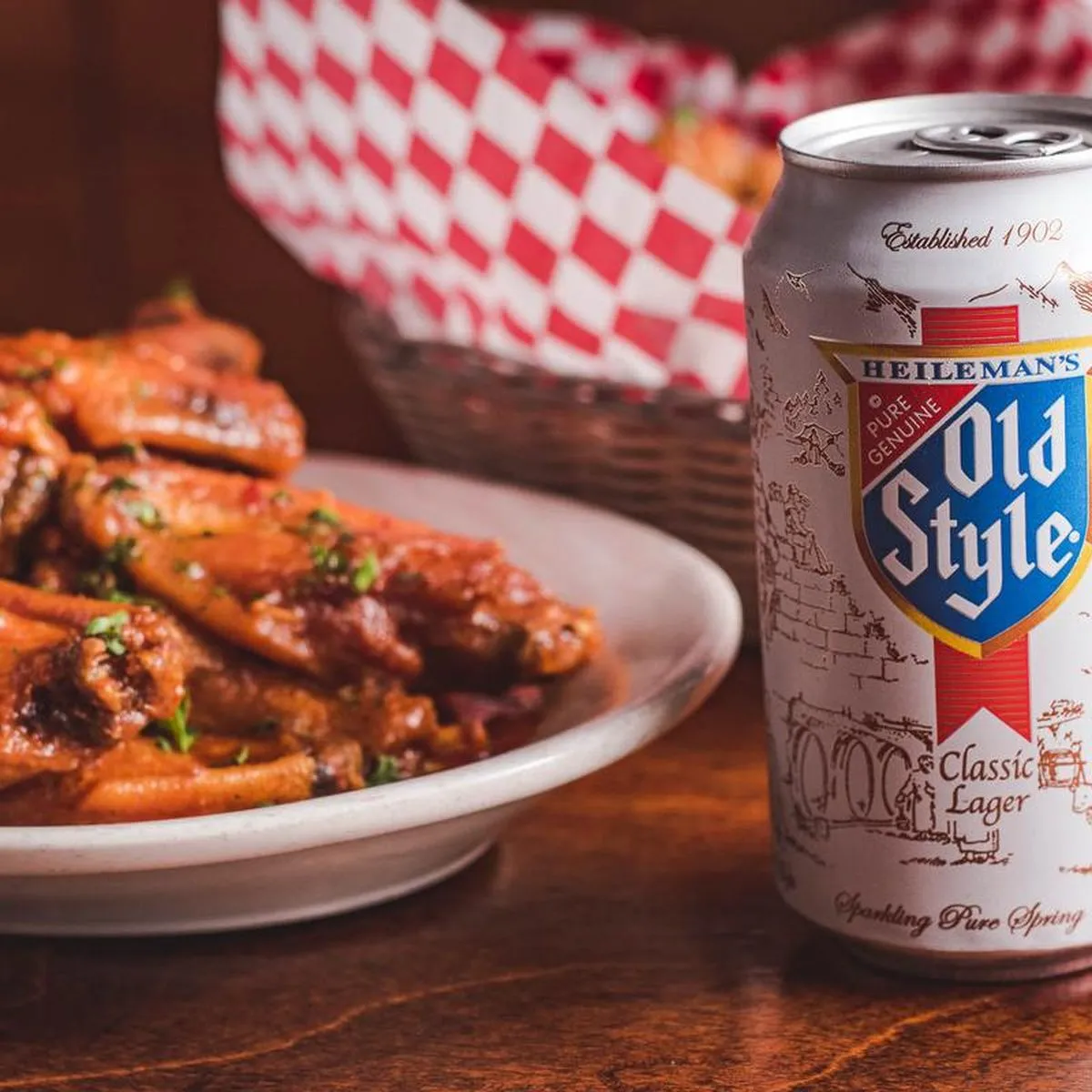 Plate of sauced chicken wings beside a can of Old Style lager, checked paper basket behind