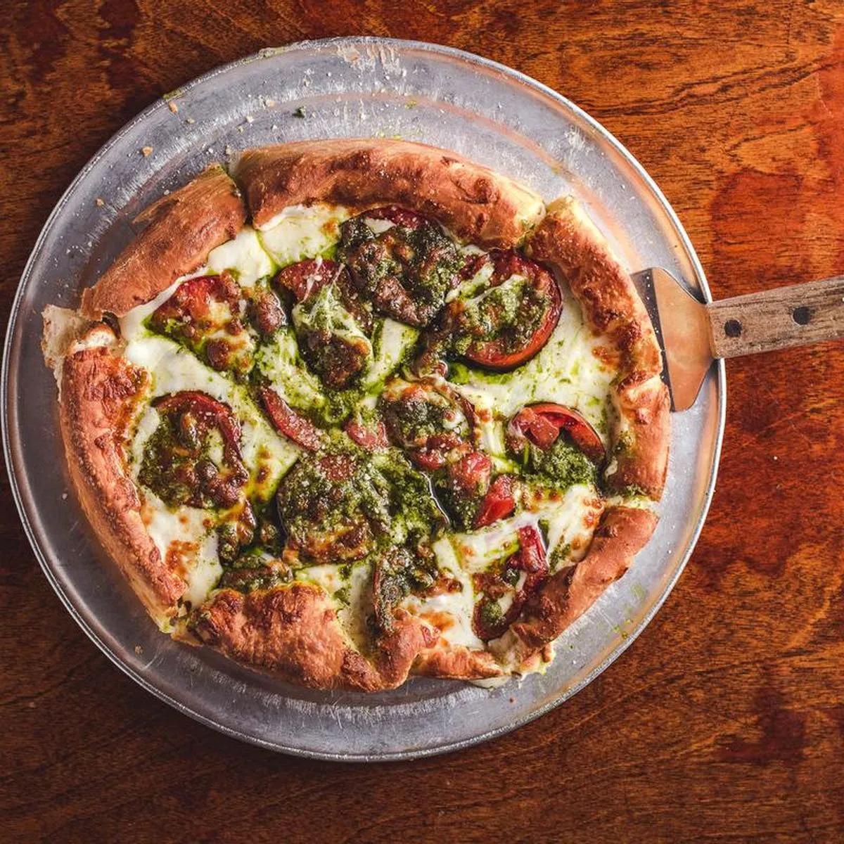 Overhead of a pesto and tomato pizza on a steel pan, the server resting on the rim